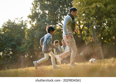 asian father and son playing soccer outdoors in park while mother and daughter watching from behind