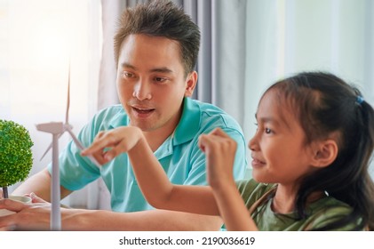 Asian Father Teaching Daughter To Play With Model Of A Windmill At The Writing Desk. Development And Learning Skills In Science And Clean Energy, Renewable Energy.