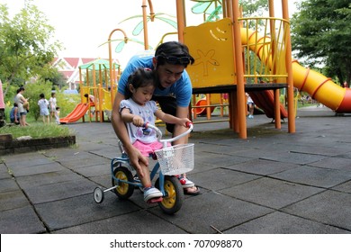 An Asian Father Is Teaching His Daughter A Bicycle At The Playground.