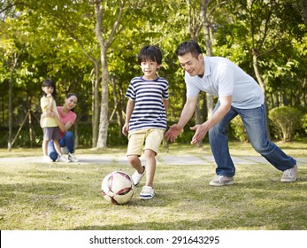 Asian Father Teaching Son To Play Soccer (football) In A Park While Mother And Daughter Watching From Behind.