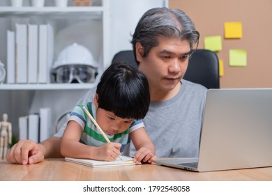 Asian Father Working At The Home Office With A Laptop Beside The Daughter Trying To Do His Homework. New Lifestyle Normal During A Quarantine. Concept Of Stay Home, Freelance And Fatherhood Concept