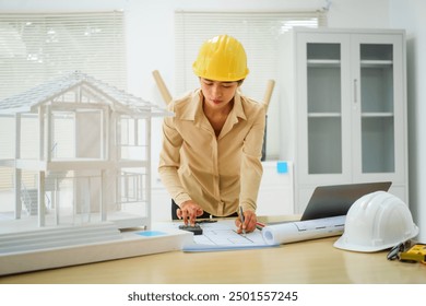 An Asian female architect analyzes blueprints at her desk in the office. She specializes in house design, focusing on roofs and walls, and collaborates with construction contractors in her company. - Powered by Shutterstock