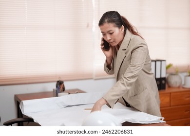 Asian female architect in beige blazer talking on phone while reviewing blueprints on desk. Professional office setting with focus on multitasking and communication in architectural planning. - Powered by Shutterstock