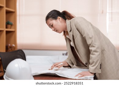 Asian female architect reviewing project plans on table in an office. Wearing beige blazer, focused on work. white safety helmet is on the table, indicating her involvement in construction projects. - Powered by Shutterstock