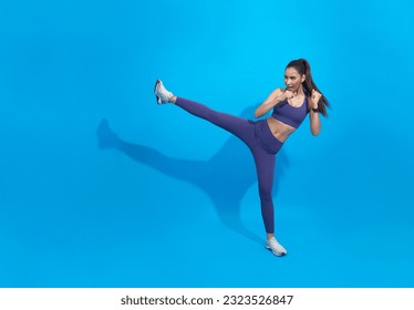Entrenamiento atleta asiática, patadas en las piernas, patadas al aire en ropa deportiva. Mujer muscular entrenada pateando con los pies elevados, ejercicios de kickboxing movimientos, fondo azul.