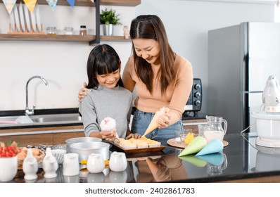 Asian female baker pastry chef mother helping teaching little girl daughter standing smiling holding piping decorating colorful whipped cream on cupcakes surface after baking tasty homemade bakery. - Powered by Shutterstock