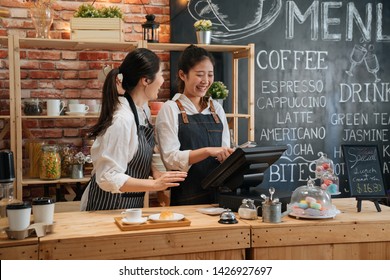 Asian Female Barista Telling Something Funny To Colleague While Working. Two Young Girls Coffeehouse Staff Laughing Together And Chatting Gossip In Modern Coffee Store. Customer Meal On Bar Counter