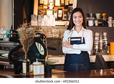 Asian Female Barista Wear Jean Apron Crossed Her Arms At Counter Bar With Smile Face, Cafe Service Concept, Owner Business Start Up.