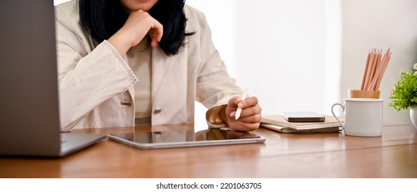 An Asian Female Boss Or Businesswoman Sits At Her Office Desk, Planning And Working On Her Project On Digital Tablet Touchpad. Cropped Image