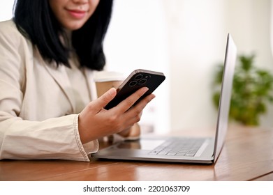 An Asian Female Boss Or Businesswoman Working At Her Office Desk, Sipping Her Morning Coffee And Using Her Smartphone. cropped Image