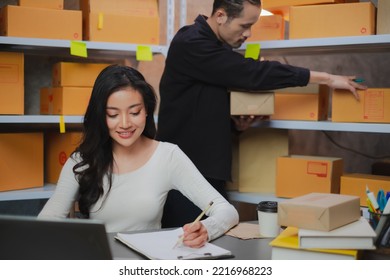 Asian Female Business Owner Or Freelance Is Looking And Write Down An Order While Her Partner Is Working With Parcel Box. Concept Of Two People, Co-worker, Small Business In Warehouse And Inventory.