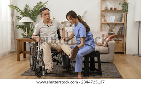 asian female care attendant helping senior man on wheelchair doing rehab training. she slowly lifts his injured leg while he shows painful face in the living room at home