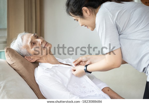 Asian female caregiver taking care of helping\
elderly patient get dressed,button on the shirt or changing clothes\
for a paralyzed person,senior woman with paralysis of limbs,body or\
muscles weakness