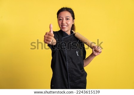 asian female chef holding wooden rolling pin with thumbs up on isolated background