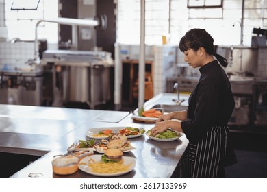 Asian female chef preparing salad in commercial kitchen with plated food and prep table, copy space. Cooking, culinary, restaurant, industrial, stainlesssteel, utensils, natural-light