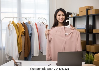 Asian Female Choosing Many Clothes On The Rail Hanging And Trying On Dress. Woman Shopping Clothes In Fashion Store. Looking For T-shirt Or Dress And Fitting Measure In Shop