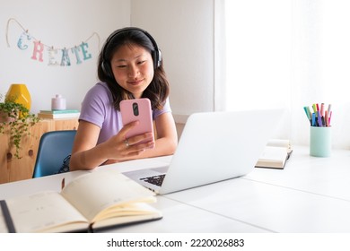 Asian female college student at home using laptop , phone and headphones while studying. Reading text message.