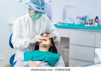 Asian Female Dentist Doctor In PPE Wearing Protective Facemask And Shield During Coronavirus Pandemic Examining Tooth For Young Girl Patient On Dental Chair For Oral Care Treatment Procedure At Clinic