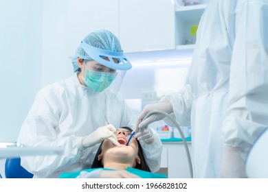 Asian Female Dentist In Lab Coat Wearing Protective Face Mask And Shield Examining Tooth For Young Girl Patient On Dental Chair. Dental Assistant Use Suction Machine For Treatment Procedures At Clinic