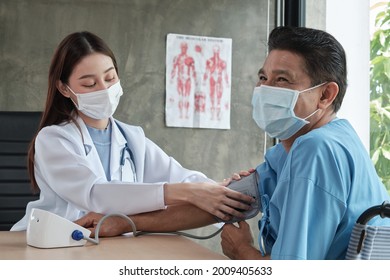 An Asian Female Doctor With Face Mask Checks The Health Of An Elderly Handicapped Man In A Wheelchair By A Blood Pressure Monitor At A Hospital Clinic. Talk Therapy Is An Examination Of Nursing.