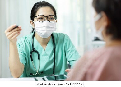 Asian Female Doctor In Green Uniform Wear Glasses And Surgical Mask Talking, Consulting And Giving Advice To Elderly Woman Patient At The Hospital. Symptoms, Disease, Flu, Sick, Covid-19, Coronavirus