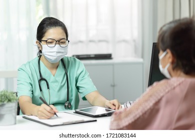 Asian Female Doctor In Green Uniform Wear Eyeglasses And Surgical Mask Writing In Checklist While Talking With Elderly Woman Patient In Medical Room At Hospital. Symptom, Disease, Flu, Covid-19