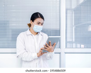 Asian Female Doctor Neatly Black Hair Wearing A Blue Medical Face Mask In A White Lab Coat Using And Looking At Digital Tablet In Hospital. Healthcare, Technology, And Medicine Concept.