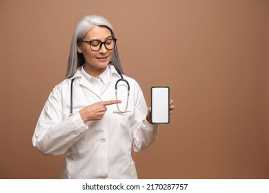 Asian Female Doctor Pediatric, Physician, Therapist Wearing White Medical Gown Points Finger On Empty Smartphone Screen, Mock Up, Copy Space, Presenting Mobile Healthcare App