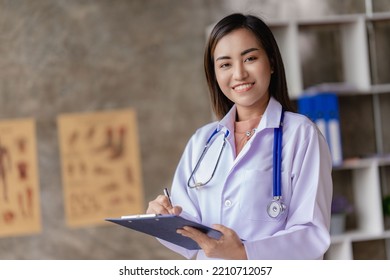 Asian Female Doctor Sitting At A Hospital Desk, Giving Health Care And Disease Prevention Advice, Convenient Online Services To Patients And Smiling, Writing Prescriptions To Order Medicine.