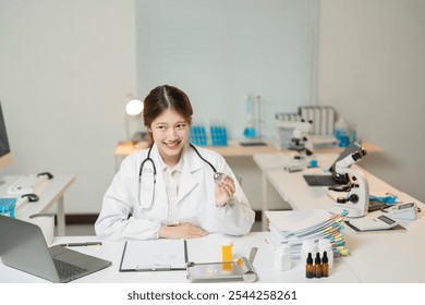 Asian female doctor smiles while holding a stethoscope in a medical laboratory, with various medical equipment and documents surrounding her - Powered by Shutterstock