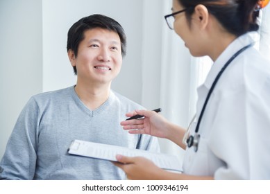 Asian Female Doctor Taking Note And Writing Information In Checklist Paper On Clipboard During Examining And Talking With Young Male Patient In Medical Room Of Hospital.