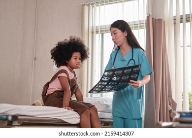 Asian Female Doctor In Uniform Health Checks African American Child, Illness Patient, Diagnosis Explains X-ray Film In Emergency Room Bed At Hospital Ward, Pediatric Clinic, Kids Examination Consult.