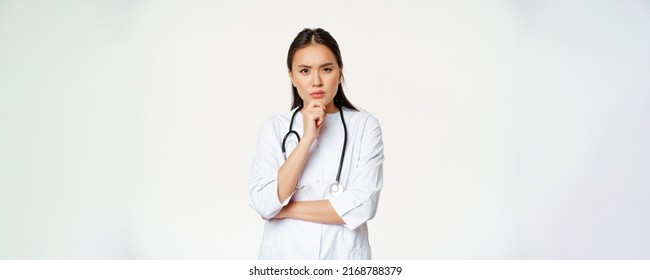 Asian female doctor in uniform thinking, looking serious and concerned, touching chin while pondering, listening patient at hospital, white background