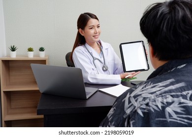 Asian Female Doctor Uses Digital Tablet To Provide Online Consultation With Patients In Hospital Examination Room.