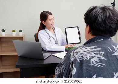 Asian Female Doctor Uses Digital Tablet To Provide Online Consultation With Patients In Hospital Examination Room.