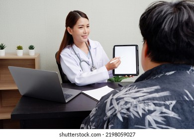 Asian Female Doctor Uses Digital Tablet To Provide Online Consultation With Patients In Hospital Examination Room.