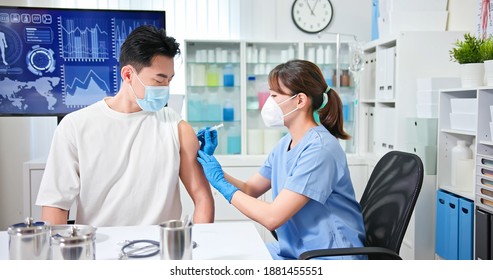 Asian Female Doctor Wearing Face Mask And Gloves Is Making A Vaccination In The Shoulder Of Male Patient In Hospital