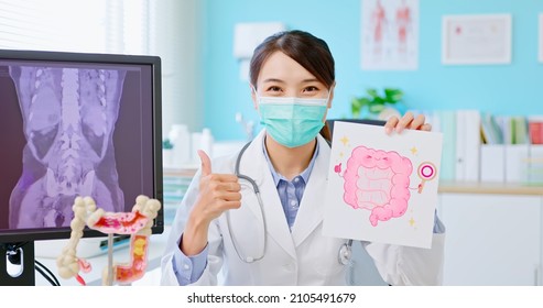 Asian Female Doctor Wearing Face Mask To Prevent COVID19 Looks At You Gives Health Education At Hospital - She Holding Intestine Picture With Colonoscopy Results And Xray On Computer In Clinic