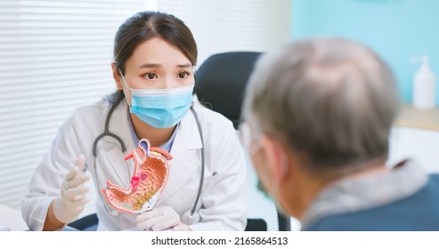 Asian Female Doctor Wearing Face Mask Is Showing Stomach Model And Explaining To Elder Senior Man Patient In Hospital