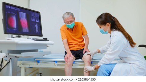 Asian Female Doctor Wearing Face Mask Is Showing X Ray Of Knee Joint On Computer Screen And Explaining To Elder Senior Man Patient In Hospital 