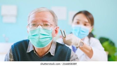 Asian Female Doctor Wearing Face Mask Is Using Tuning Fork For Hearing Tests To Elder Senior Man Patient In Hospital