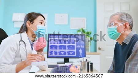 asian female doctor wearing face mask is showing a model of brain and explaining to elder senior man patient in hospital