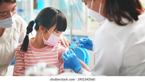 Asian Female Doctor Wearing Gloves And Isolation Mask Is Making A COVID-19 Vaccination In The Shoulder Of Child Patient With Her Mother At Hospital