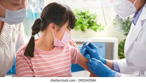 Asian Female Doctor Wearing Gloves And Isolation Mask Is Making A COVID-19 Vaccination In The Shoulder Of Child Patient With Her Mother At Hospital