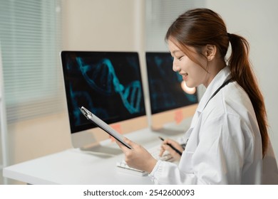 Asian female doctor wearing lab coat and stethoscope is analyzing a dna double helix structure on her computer screen. Taking notes on a clipboard and smiling. In a modern medical research laboratory - Powered by Shutterstock