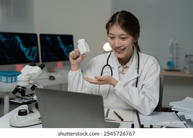 Asian female doctor wearing lab coat and stethoscope showing medicine bottle during video conference on laptop in laboratory with microscope and dna double helix on computer screens - Powered by Shutterstock