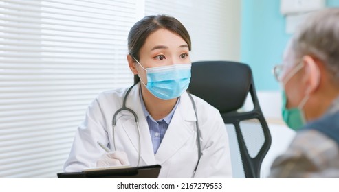 Asian Female Doctor Wearing White Coat And Face Mask Is Diagnosing Gray Hair Elder Senior Sick Male Patient In Hospital - She Takes Note On Paper Document