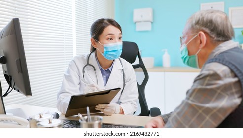 asian female doctor wearing white coat and face mask is diagnosing gray hair elder senior sick male patient in hospital - she takes note on paper document