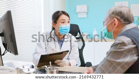 asian female doctor wearing white coat and face mask is diagnosing gray hair elder senior sick male patient in hospital - she takes note on paper document