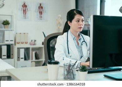 Asian Female Doctor Working With Personal Computer And Writing On Paperwork. Hospital Background. Chinese Woman Medical Staff Wear White Robe In Clinic Office Typing Online Patient Document Internet.